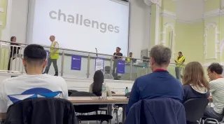 Louie Christie on a stage holding a microphone. A screen behind him says 'challenges'. Several people are on stage
            laughing at something. Audience of geeks in the foreground.
