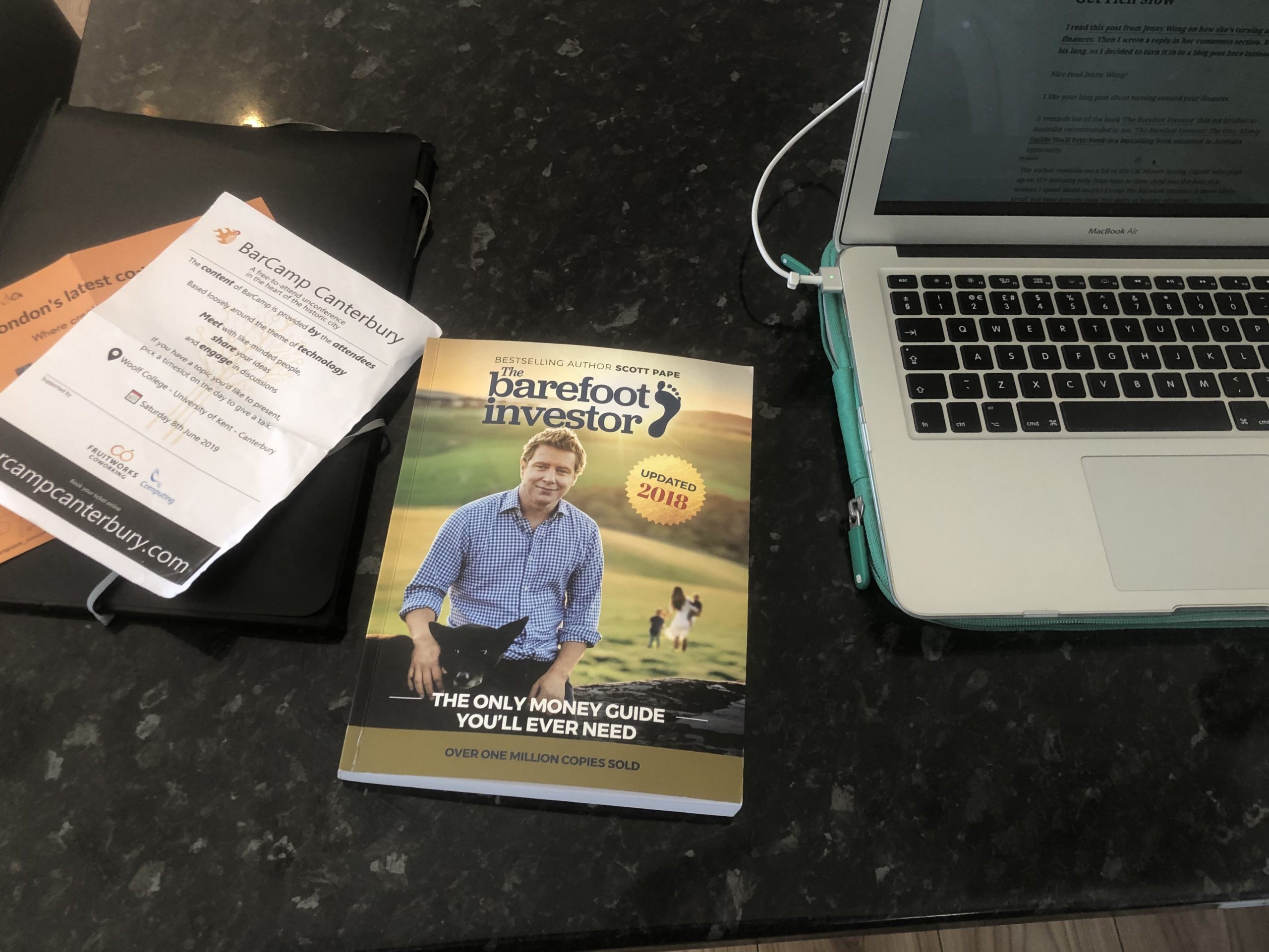 A table with The Barefoot Investor book on it, and also an Apple Mac Laptop and flyers for a BarCamp and a co-working space.