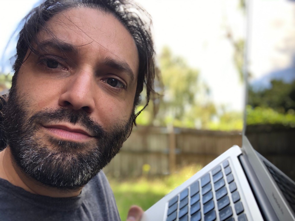 Louie Christie outdoors on a sunny day in a garden holding a mac laptop up. Long hair is windswept across his gormless face.