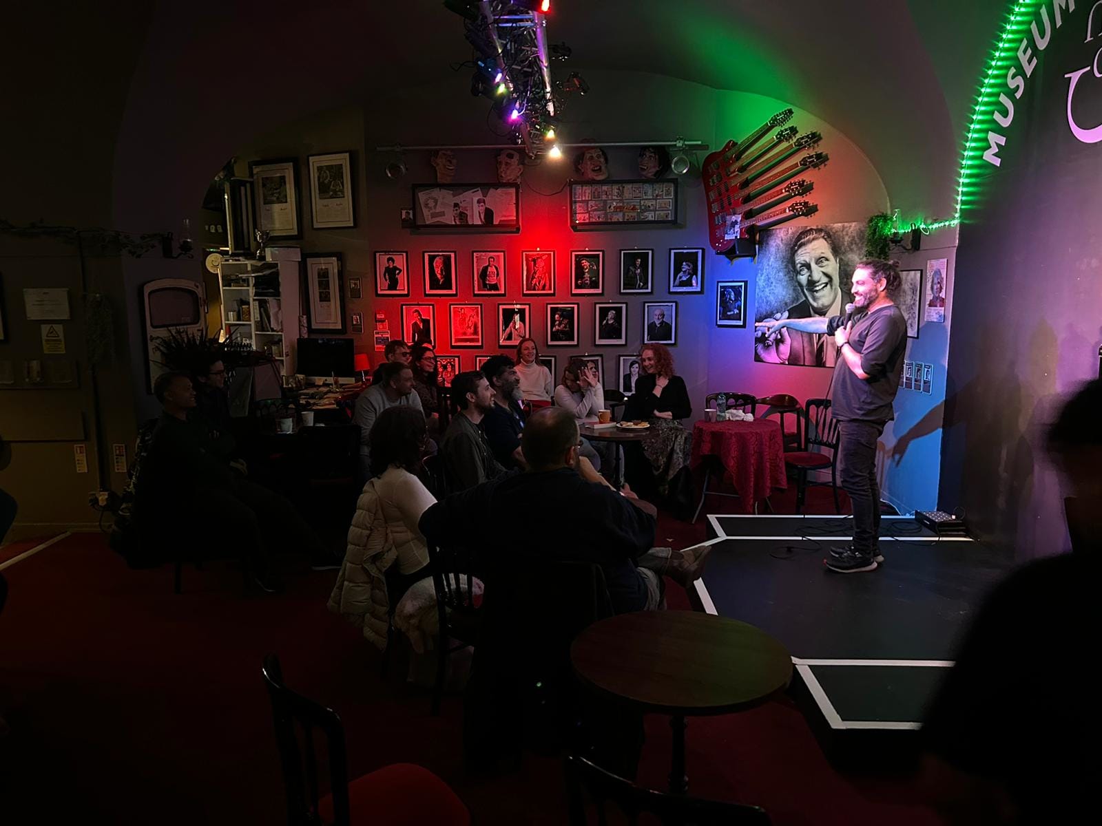 Louie Christie performing stand-up comedy while audience laughs. On the wall behind there is a big painting of Tommy Cooper and smaller photos of various famous stand up comedians. Louie is pointing at the audience and seems to be having a good time. His hair is in a man bun. He's pointing at a woman in the audience and she has her hands crossed in front of her face as if she doesn't want to talk. Which is ironic, because it was about the only time in the whole night that she shut up.