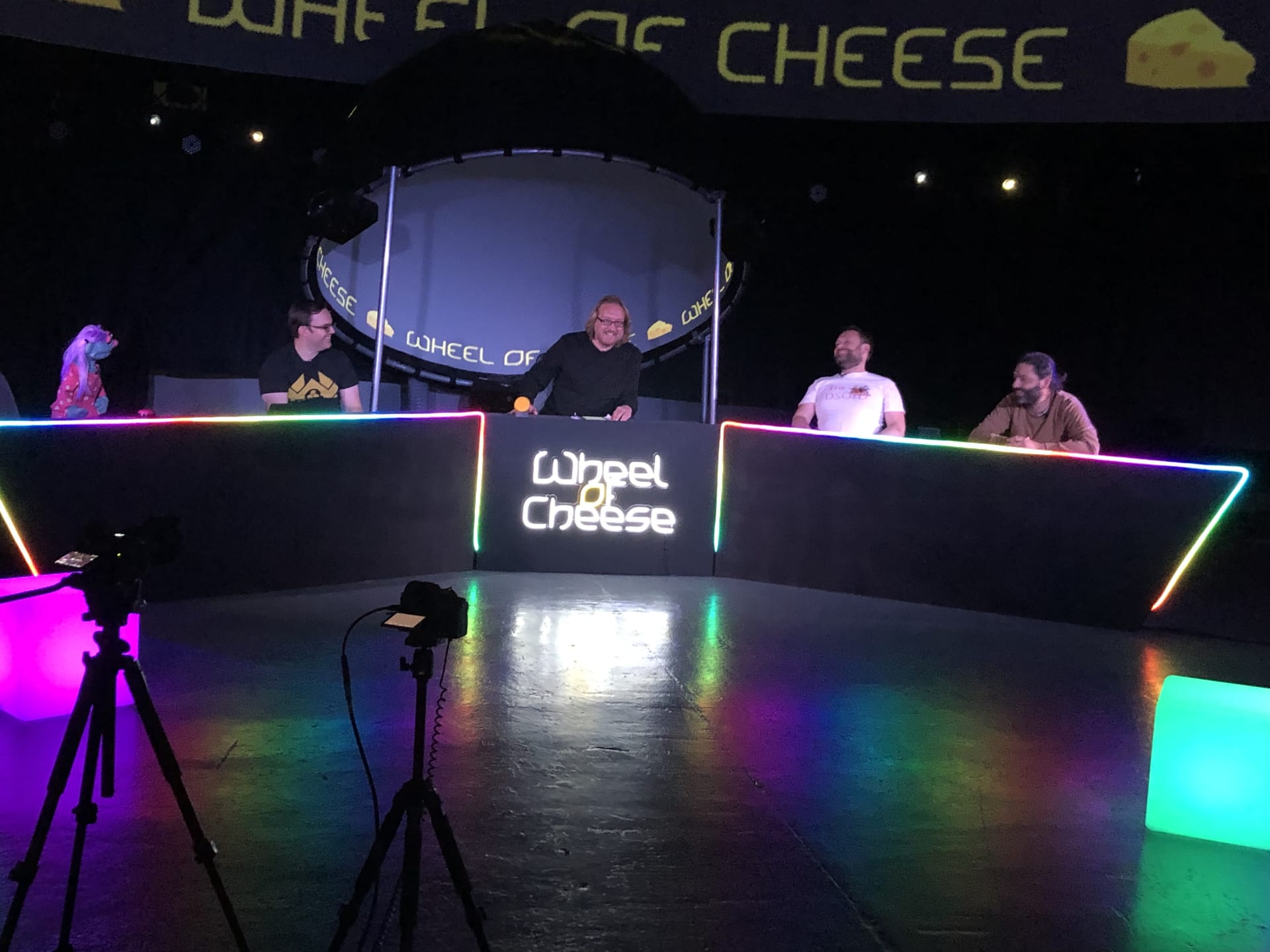 Panel quiz show studio. A host and four contestants laughing. Louie Christie is on the far right. They are all sat behind a long desk that forms part of a colourful colourful set. A neon sign reads 'Wheel of Cheese'.