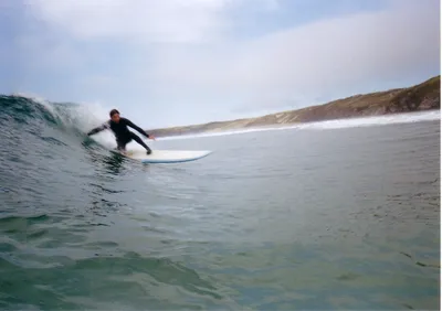 Louie Christie surfing photo A surfer on a wave. He's crouching, touching the wave with his hand as the wave breaks. The wave is chest high,
blue-green color. There are brown-green hills in the background on the land.